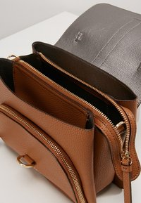 Borsa a tracolla in pelle marrone con texture, dotata di numerosi scomparti con zip e hardware dorato, abbinata a un flap in pelle liscia grigia.