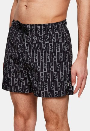 Hombre vistiendo shorts de baño negros con patrón EA7 y cintura con cordón, mostrando torso, piernas y mano derecha contra un fondo claro.