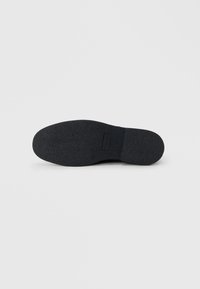 Suela de zapato de goma negra con superficie texturizada, forma redondeada y un ligero tacón. Presenta una pequeña etiqueta rectangular cerca del arco.