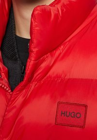 Červená péřová bunda s vysokým límcem, na které je umístěna nášivka HUGO. Materiál je lesklý a hladký, s prošívanou strukturou a zapínáním na zip.