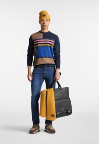 Maglione multicolore con strisce orizzontali, jeans scuri, berretto di lana giallo, tiene una borsa nera e una sciarpa. Sneakers con dettagli marroni.