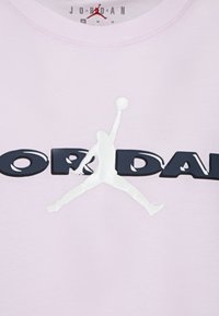 Svetlo roza majica s kratkimi rokavi in okroglim izrezom, ki ima bel logotip "Jumpman" in izrazite mornarsko modre črke, ki črtijo "JORDAN" spredaj.
