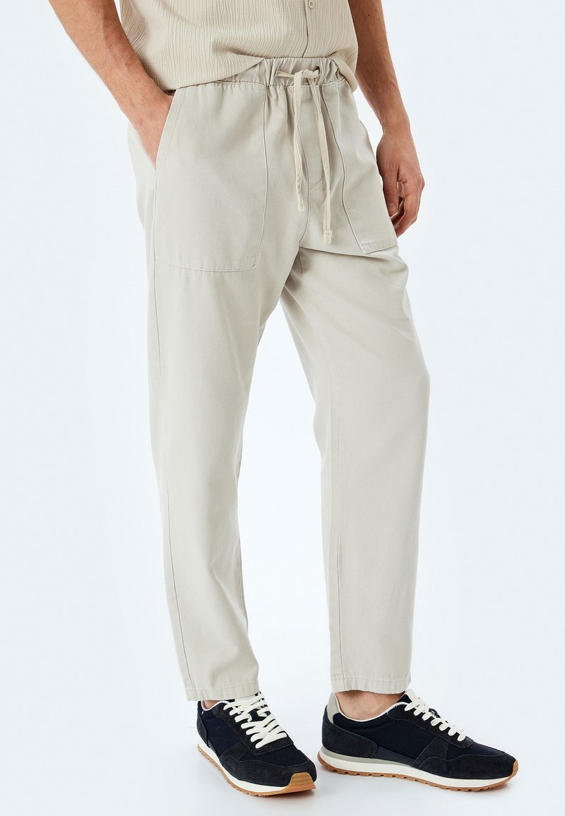 Pantaloni elasticizzati color beige chiaro con vita regolabile, dotati di tasche laterali e caviglie affusolate. Abbinati a sneakers scure con lacci bianchi.