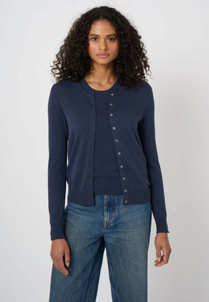 Femme aux cheveux bouclés portant un cardigan bleu marine boutonné par-dessus un haut assorti et un jean bleu, debout devant un fond uni.
