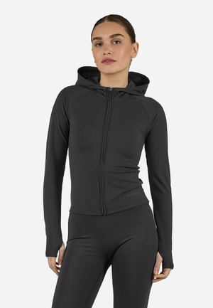 Γυναίκα που φορεί εφαρμοστό μαύρο φούτερ με φερμουάρ και τρύπες για τους αντίχειρες, μαζί με μαύρο κολάν, σε απλό φόντο.