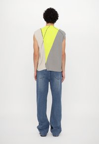 Personne aux cheveux bouclés portant un haut en tricot sans manches coloré en beige, jaune fluo et gris, associé à un jean bleu ample, se tenant de dos.