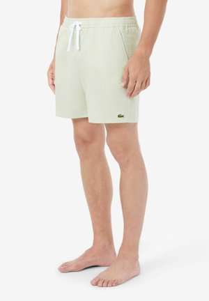 Mann trägt hellgrüne gestreifte Badeshorts mit weißem Kordelzug und kleinem grünem Krokodil-Logo, steht barfuß vor weißem Hintergrund.