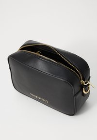 Sac en cuir noir à porter croisé avec fermeture éclair, quincaillerie dorée, bords arrondis et nom de la marque embossé sur le devant. Design compact et rectangulaire.