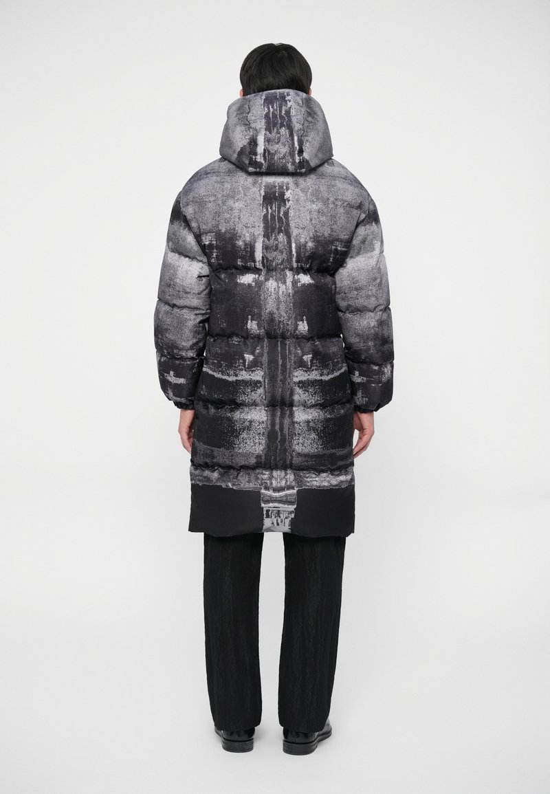 Pufferjakke i grå nuancer med et tekstureret print, forsynet med hætte og langt design. Sammen med sorte bukser og sko.