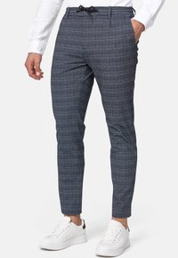 Pantalons ajustés à carreaux bleus avec taille élastique et poches latérales, associés à des baskets blanches, mettant en avant un design moderne et sur mesure.
