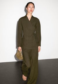 Olijfgroene linnen wikkeljas en bijpassende wijde broek. Model houdt een geweven handtas vast en draagt sandalen. Minimalistisch ontwerp en textuur.