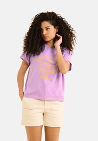 T-shirt en coton lilas avec des manches courtes retroussées et des graphiques jaune vif représentant un verre à cocktail et un design de soleil, associé à un short beige.