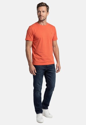 Man met een oranje T-shirt met korte mouwen, donkerblauwe spijkerbroek en witte sneakers, staand tegen een effen lichte achtergrond.