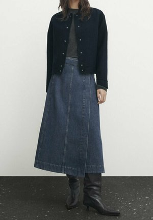 Femme portant une veste sombre boutonnée, un haut gris foncé, une longue jupe en jean et des bottes noires à talons mi-mollet, debout sur un sol sombre.