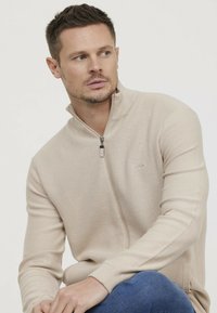 Sweater beige zippé avec un motif en maille texturée, manches longues et col haut. Associé à un jean bleu, mettant en avant un look décontracté.