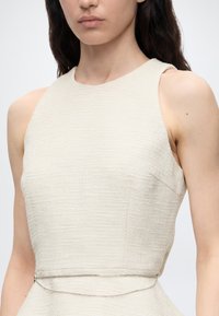 Femme portant une robe crème texturée et sans manches avec une ceinture chaîne argentée, montrée des épaules à la taille sur fond uni.