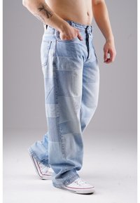 Hellblaue Patchwork-Jeans mit unterschiedlichen Denim-Strukturen und einer lockeren Passform, ergänzt durch weiße Leinensneaker mit roten Akzenten.