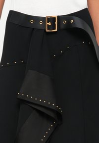 Melna svārku ar plūstošu dizainu, kas aprīkota ar elegantu jostu ar zelta sprādzi un zelta kniedēm pa apakšmalu un auduma malām.