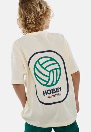 T-shirt crème avec un grand motif de volleyball vert et blanc et le texte "HOBBY depuis 1986" au dos. Manches courtes, coupe décontractée.