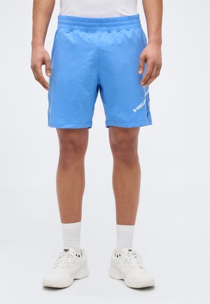 Moški, ki nosi svetlo modre atletske kratke hlače z logotipom »HEAD« na levi nogi, bele posadke nogavice in bele superge, stoji ob enobarvni ozadju.