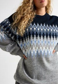 Donna con capelli biondi ricci che indossa un maglione lavorato a maglia con un motivo grigio, blu e nero, con le mani sui fianchi, su uno sfondo neutro.