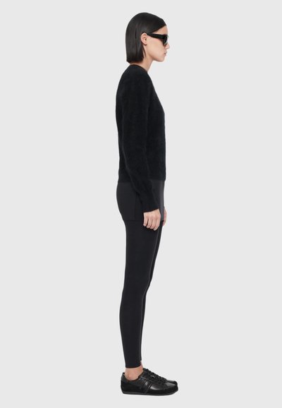 Une femme se tient de profil, portant des lunettes de soleil foncées, un pull noir, un pantalon ajusté et des baskets noires sur un fond gris uni.