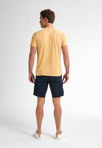 Pánské žluté bavlněné tričko, námořnické cargo šortky a světlé tenisky. Tričko má kulatý výstřih; šortky mají postranní cargo kapsy.