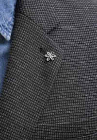 Dunkel gemusterter Blazer aus strukturiertem grauem Stoff, mit einer silbernen Blumen-Anstecknadel und einem blauen Jeanshemd darunter.