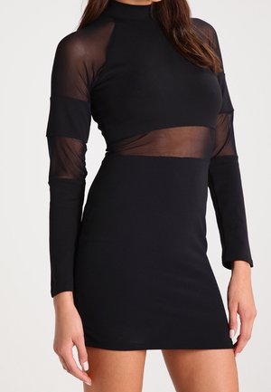 Femme portant une mini robe noire ajustée avec des panneaux en maille transparente sur les manches et le ventre, debout devant un fond uni.