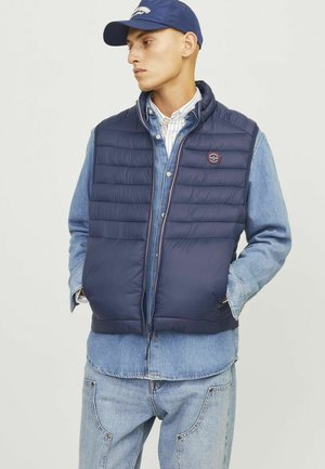 Navy puffer vest with horizontal quilted texture, front zipper, side pockets, and logo patch. Worn over a denim shirt and light jeans.