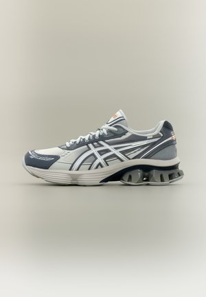 Chaussure de sport grise et blanche avec un design de talon unique en forme de ressort et logo Asics sur le côté, présentée de profil sur un fond neutre.