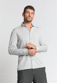 Helles graues Langarmhemd mit Button-down-Kragen, das eine weiche Textur, eine Knopfleiste vorne und passende Manschetten aufweist. Getragen mit dunkler Hose.