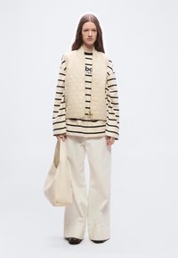 Γυναίκα με μπεζ καπιτονέ γιλέκο πάνω από ριγέ μακρυμάνικο πουκάμισο, φαρδιά κρεμ παντελόνια, κρατώντας τσάντα tote σε ταιριαστό μπεζ χρώμα, όρθια.