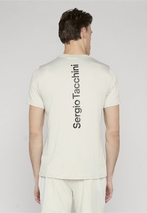 Mand set bagfra iført lysebeige t-shirt og shorts med "Sergio Tacchini" trykt vertikalt i sort på bagsiden af skjorten.