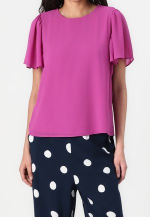 Femme portant un chemisier rose vif à manches volantes et un pantalon bleu marine à gros pois blancs.