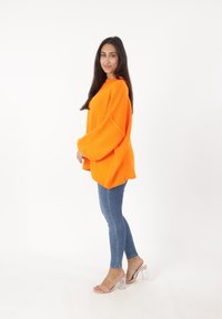 Übergroßer oranger Strickpullover mit weitem Ärmel, kombiniert mit schmalen blauen Jeans und transparenten High-Heel-Sandalen. Schlichter weißer Hintergrund.