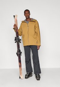 Strój narciarski z żółtą wodoodporną kurtką, szarym wewnętrznym layerem, ciemnoszarymi spodniami i czarnymi butami narciarskimi, trzymający narty z wiązaniami.