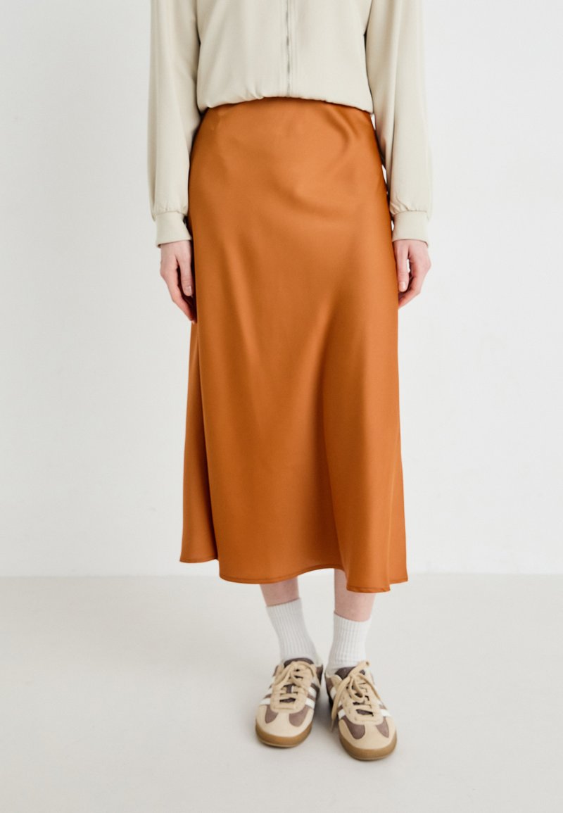 Brun sateng midi skjørt med en glatt tekstur, som har en flowy silhuett som strekker seg til midt på leggen, kombinert med beige sneakers og hvite sokker.