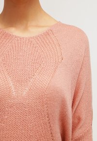 Maglione lavorato a maglia rosa chiaro con una vestibilità ampia, scollatura larga e motivi testurizzati nella zona del petto, con dettagli leggermente traslucidi.
