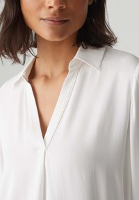 Weiße Bluse mit V-Ausschnitt, langen Ärmeln und einem glatten, leichten Stoff. Verfügt über einen klassischen Kragen und subtile Drapierungen.