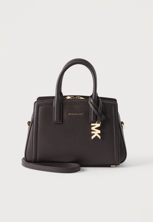 Sac à main en cuir noir avec poignées supérieures, fermeture éclair dorée et breloque logo MK dorée sur le devant, reposant sur une surface blanche.