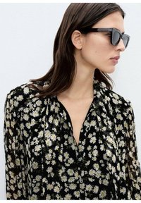 Svart blus med blommönster av vita prästkragar, knäppning framtill och lätt tyg. Modellen bär mörka solglasögon.