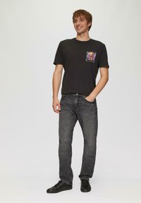 Schwarzes Baumwoll-T-Shirt mit kurzen Ärmeln, ausgestattet mit einem bunten grafischen Taschenaufdruck, kombiniert mit dunkelgrauen Jeans und schwarzen Turnschuhen.