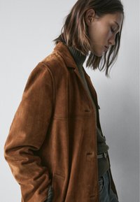 Bruine suède jas met een ontspannen pasvorm, met een kraag, knopen aan de voorkant en zijzakken. Draagt over een textuurrijke groene trui.