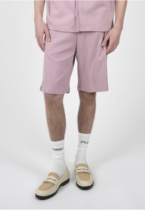 Unterkörper einer Person, die hellviolette Shorts, weiße Markensocken und beigefarbene Loafers mit strukturierten Details auf einem einfarbigen Hintergrund trägt.