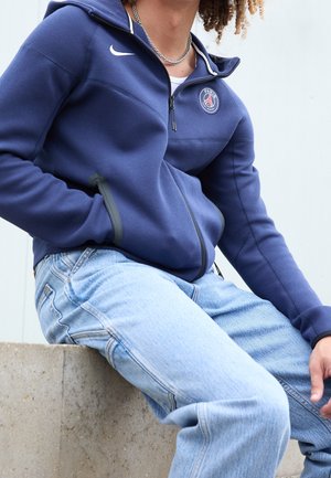 Mann trägt einen marineblauen Paris Saint-Germain Zip-Hoodie und hellblaue Jeans, sitzt auf einer Betonkante vor einem schlichten Hintergrund.