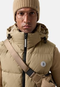 Beige puffervest med høj krave, sort lynlås og logoaccent; sammen med en lysebrun ribbet hue og minimalistiske briller.