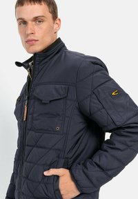 Mørkeblå quiltet jakke med høj krave, lynlås lukning og to forlommer. Har diskret syning og et logo på ærmet.