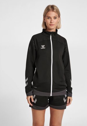 Zwarte sportjack met witte ritssluiting, logo en zilveren accenten. Heeft geperst mouwen en een getailleerd ontwerp. Gepaarde met zwarte shorts.