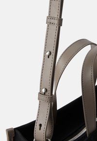 Bolso de tela negra con correas de cuero marrón. Las características incluyen longitud de correa ajustable y acentos de hardware plateado.